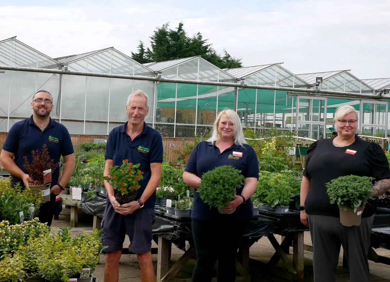 Grimsby Garden Centre Team holding plants with a set of greenhouses in the background