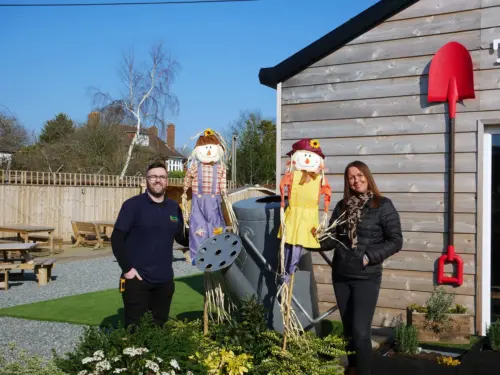 Man and woman holding scarecrows outside near oversized spade and watering can
