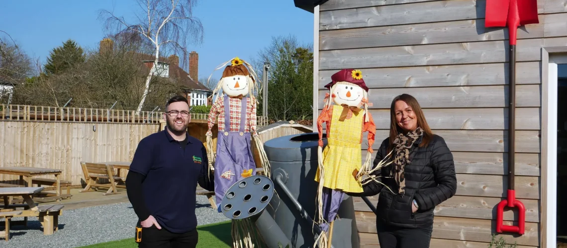 Man and woman holding scarecrows outside near oversized spade and watering can
